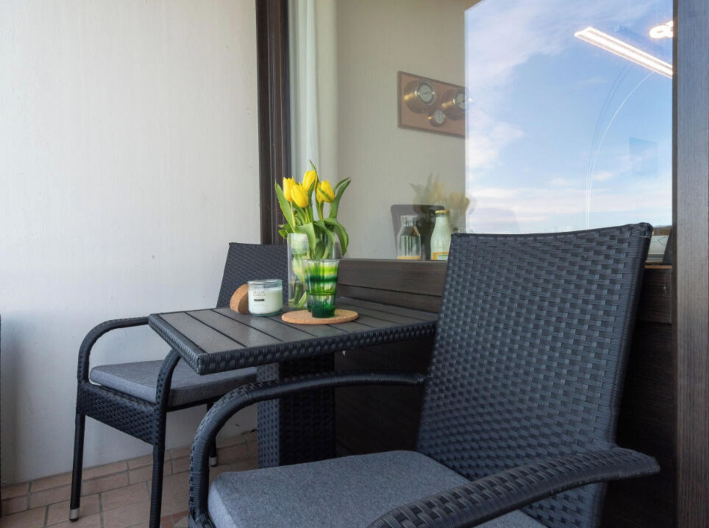 Een balkon met een tafel, twee stoelen, een vaas met gele bloemen en een klok aan de muur.