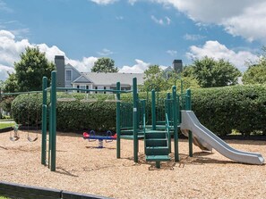A playground with a slide and swings, surrounded by a hedge and a building in the background. - Landing | Incredible 1BD, Clubhouse, Gym (Murfreesboro)