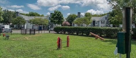 A grassy outdoor area with a playground, a trash can, and a green trash bin.