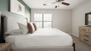 Ein Schlafzimmer mit einem Bett, einem Deckenventilator, einem Fenster und einem Fernseher auf einer Holzkommode.