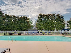 Ein Schwimmbecken mit Liegestühlen und einem Zaun, umgeben von Bäumen und einem Gebäude im Hintergrund.
