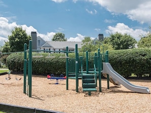 A playground with a slide and swings, surrounded by a hedge and a building in the background. - Landing | Stylish 1BD, Gym, Clubhouse (Murfreesboro)