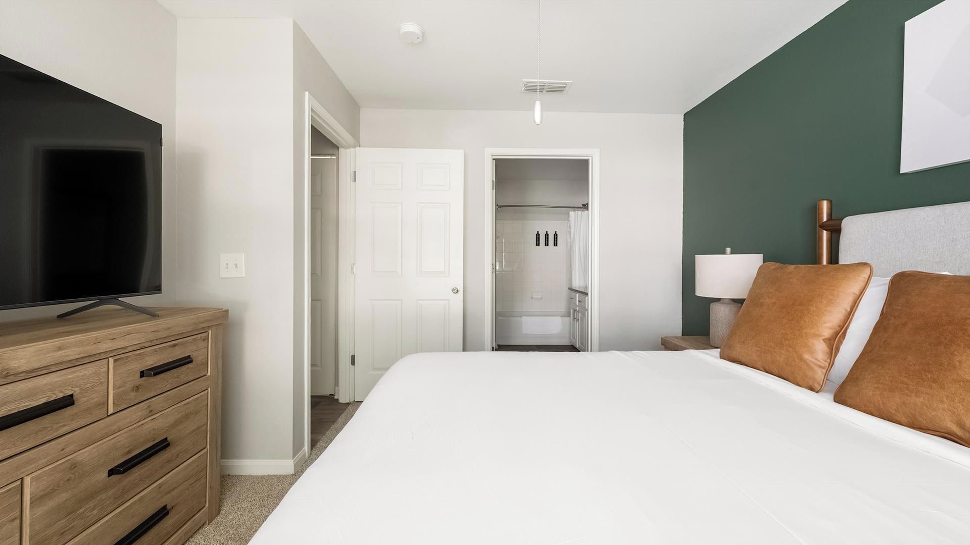 Una habitación de hotel con una cama, un televisor sobre una cómoda de madera, una mesita de noche con una lámpara y un baño visible a través de una puerta abierta.