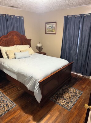 A bedroom with a wooden bed, a nightstand with a lamp, a framed picture on the wall, and a patterned rug.