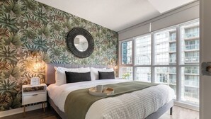 Une chambre avec un grand lit, une table de chevet, un miroir fixé au mur et une fenêtre donnant sur la ville.