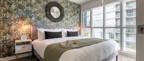 Quarto com cama grande, mesa de cabeceira, espelho fixado na parede e janela com vista para a cidade.