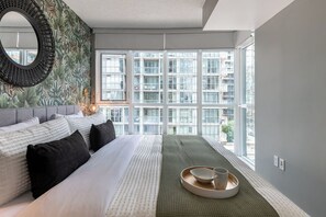 A modern bedroom with a large bed, a view of a cityscape through floor-to-ceiling windows, and a decorative mirror.