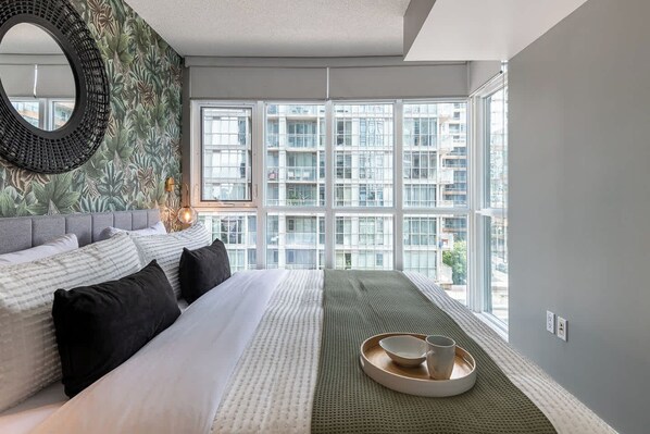 Une chambre moderne avec un grand lit, une vue sur le paysage urbain grâce à de grandes fenêtres du sol au plafond, et un miroir décoratif.