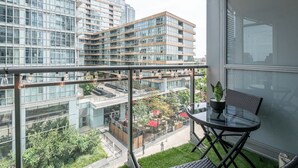 Un balcon doté d’une balustrade en verre, d’une table et d’une chaise, offrant une vue sur un paysage urbain avec des bâtiments modernes et un parc.