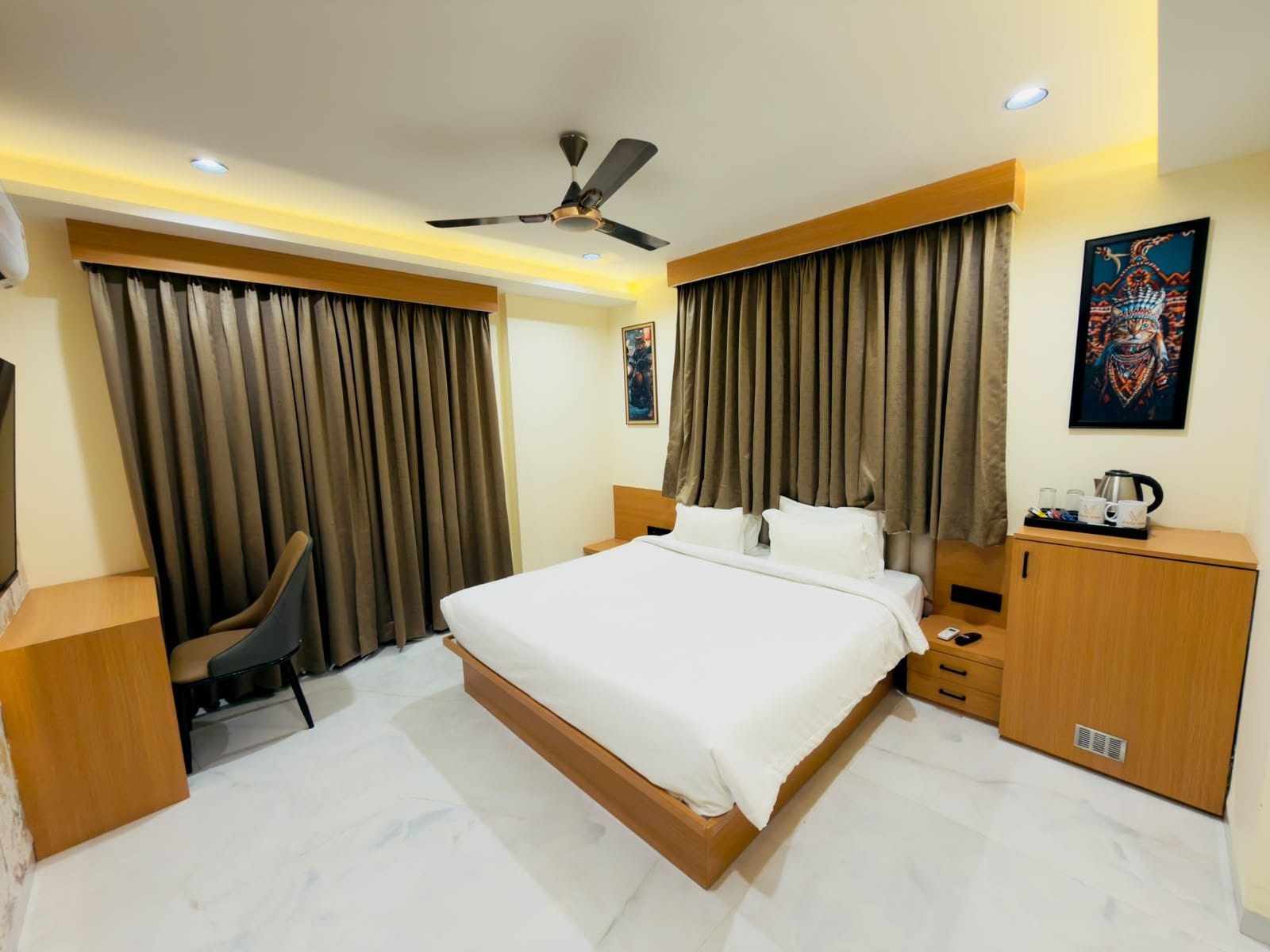 A hotel room with a bed, a desk, a chair, a wardrobe, a ceiling fan, and a painting on the wall.