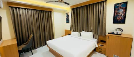 A hotel room with a bed, a desk, a chair, a wardrobe, a ceiling fan, and a painting on the wall.