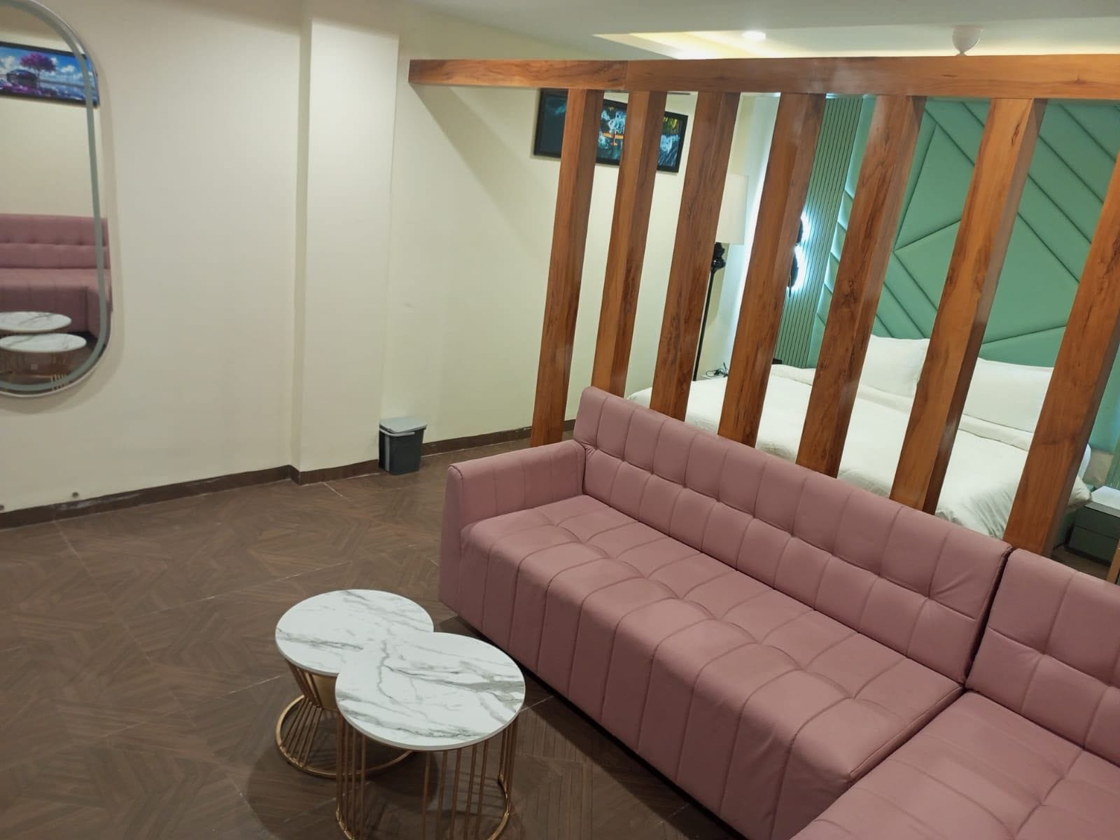A modern living room with a pink sectional sofa, two marble-topped side tables, a wooden ceiling structure, and a bed with green headboard in the background.