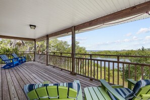 A covered outdoor deck with wooden railings, green and blue Adirondack chairs, and a view of a body of water and greenery.