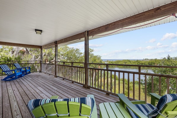 Una terraza techada al aire libre con barandas de madera, sillas Adirondack verdes y azules, y vistas a una masa de agua y follaje.