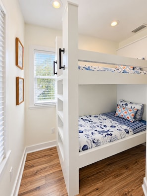 A bunk bed with a patterned bedspread, a window with blinds, and a white wall with two framed pictures.