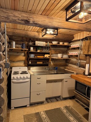 Une cuisine rustique aux murs en bois, avec un poêle blanc et des étagères remplies d’assiettes et de verres.