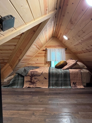 Une chambre à coucher aménagée dans un espace sous les toits, avec un plafond en pente, un lit recouvert d’une courtepointe et une fenêtre ornée d’un rideau léger.