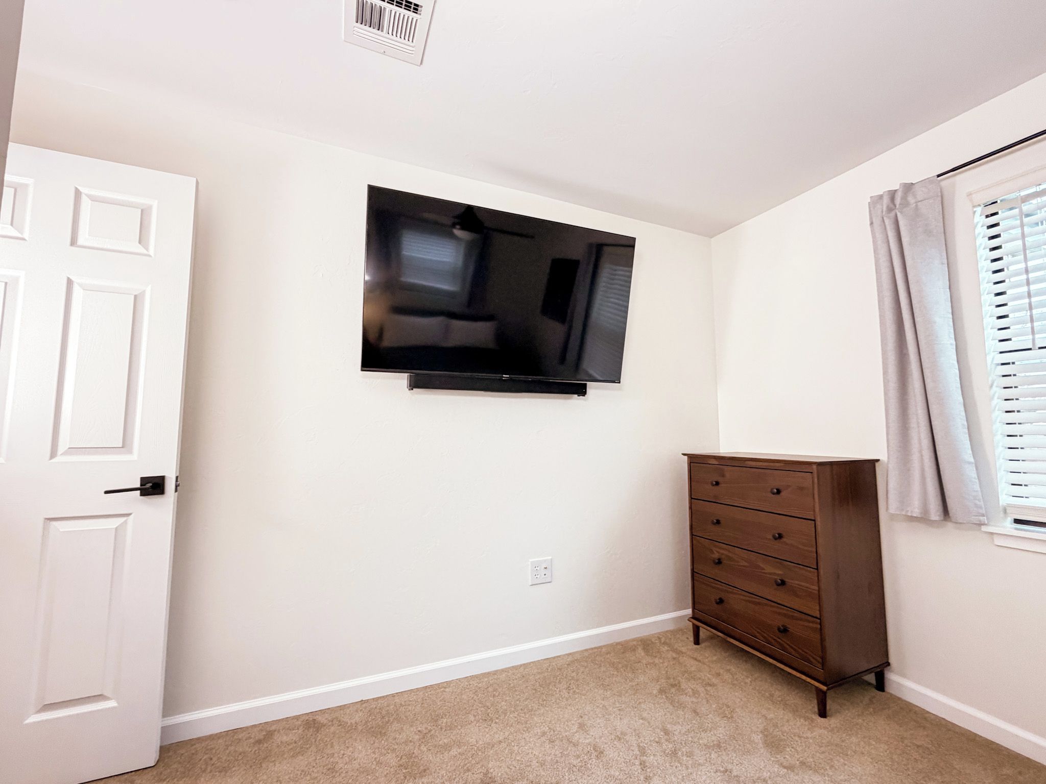 Une pièce avec une porte blanche, un téléviseur à écran plat fixé au mur, une commode en bois et une fenêtre avec des stores.