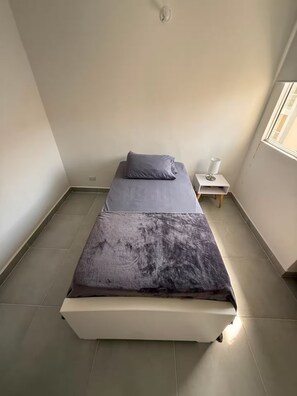 A single bed with a grey mattress and pillows, a bedside table with a lamp, and a window with a view of buildings. - Welcome to MA Cartagena • Playa • Piscina (Bolívar)
