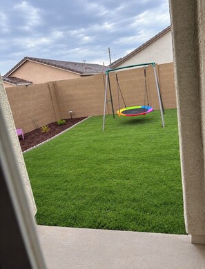 Un patio con césped artificial, un conjunto de columpios y una pared de ladrillo.