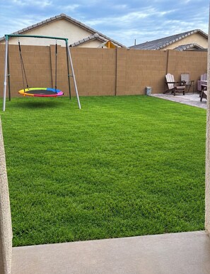 Un patio trasero con un columpio, un trampolín y un área de patio con sillas.