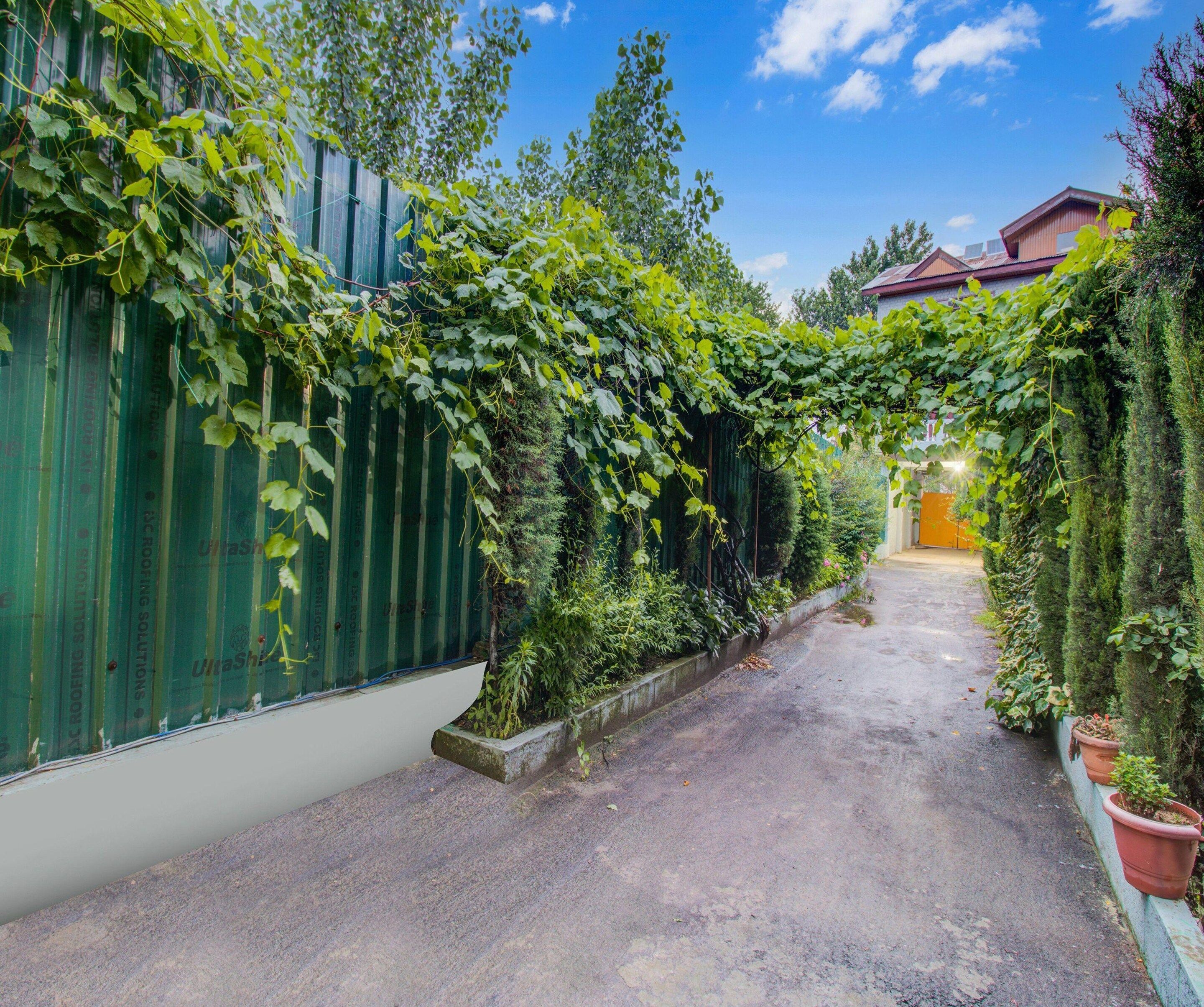 A narrow alleyway with a green metal fence covered in foliage, leading to a building with a red roof.