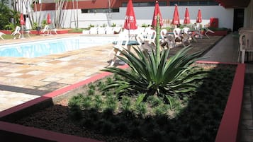 Uma área de piscina com espreguiçadeiras e guarda-sóis, rodeada por um jardim com plantas e uma palmeira.