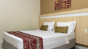 Habitació d'hotel amb llit, roba de llit blanca, una manta daurada i vermella, i un capçal de llit de fusta.