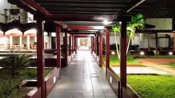 A covered walkway with a tiled floor, flanked by greenery and palm trees, leading to a building with multiple windows.