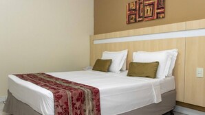 Habitación de hotel con cama, ropa de cama blanca, una manta roja y dorada, y cabecero de madera.