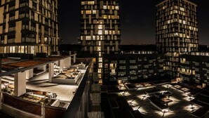 Complejo de edificios altos por la noche con ventanas iluminadas y una terraza en la azotea con mesas al aire libre.