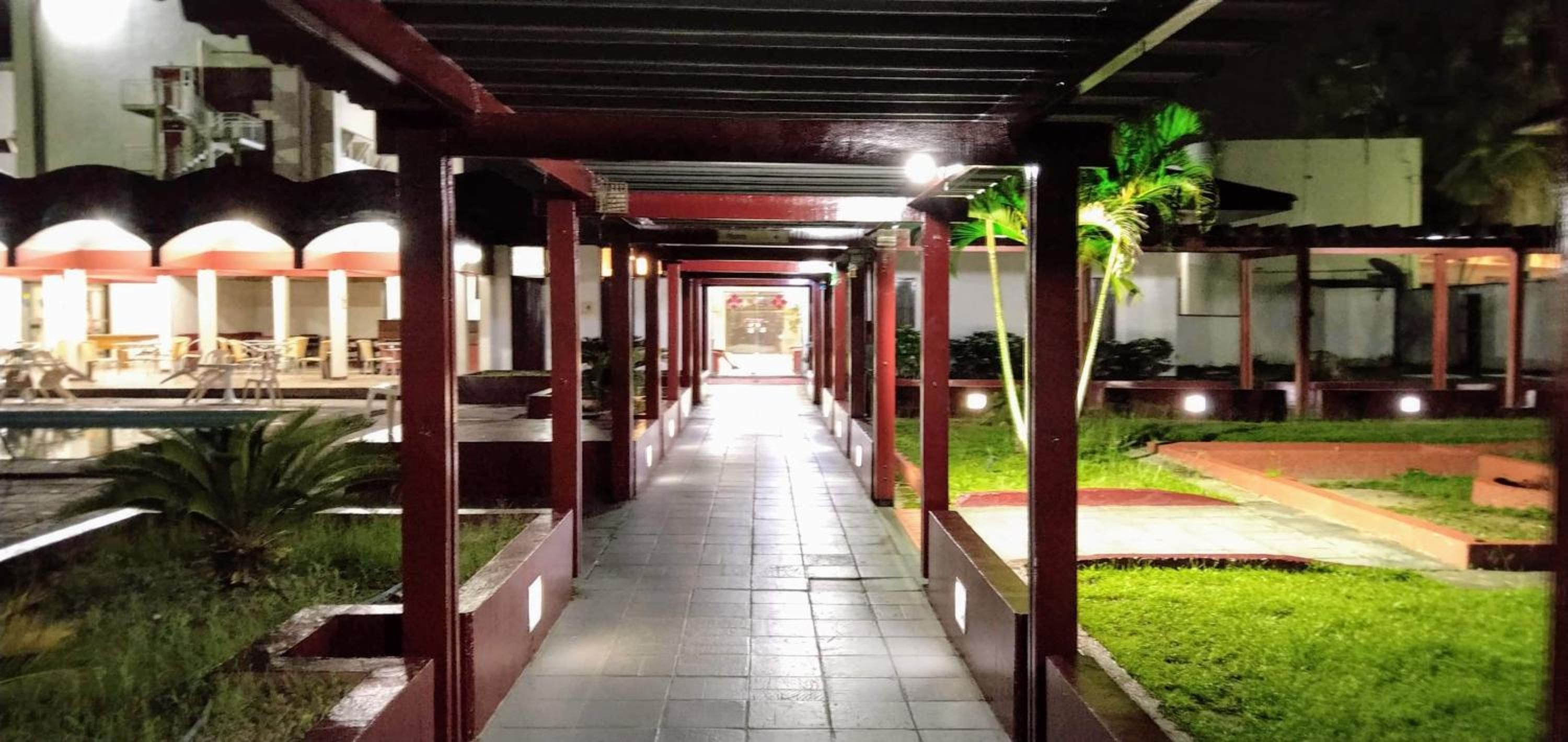 A covered walkway with a tiled floor, flanked by greenery and palm trees, leading to a building with multiple windows.