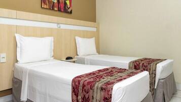 Quarto de hotel com duas camas de solteiro, cada uma com roupa de cama branca e colcha vermelha florida. Possui cabeceira de madeira, mesa de cabeceira pequena e uma obra de arte pendurada na parede acima das camas.
