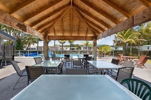 Un patio techado al aire libre con mesas y sillas, una piscina al fondo y palmeras.