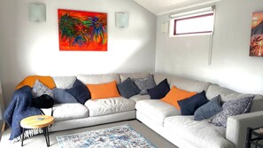 A living room with a grey sofa, multiple cushions, a small round table, a patterned rug, and two wall-mounted light fixtures.