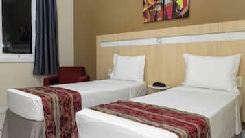 Quarto de hotel com duas camas de solteiro, uma cadeira vermelha, uma mesa pequena e uma janela com cortinas.