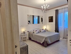 Een slaapkamer met een bed, nachtkastje, spiegel, kroonluchter en gordijnen.