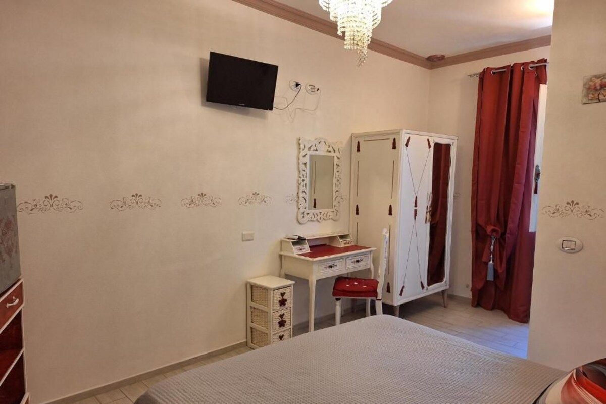 A bedroom with a bed, a television mounted on the wall, a wardrobe, a desk with a chair, and a red curtain.