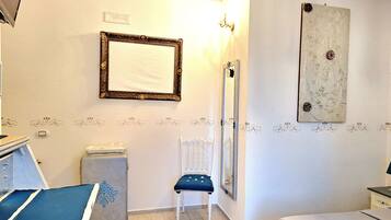 Una habitación con cama, una silla, un escritorio con computadora, un espejo y una pared con detalles decorativos.