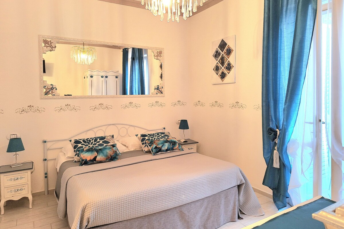 Een slaapkamer met een bed, nachtkastjes, een spiegel, een kroonluchter en een raam met gordijnen.