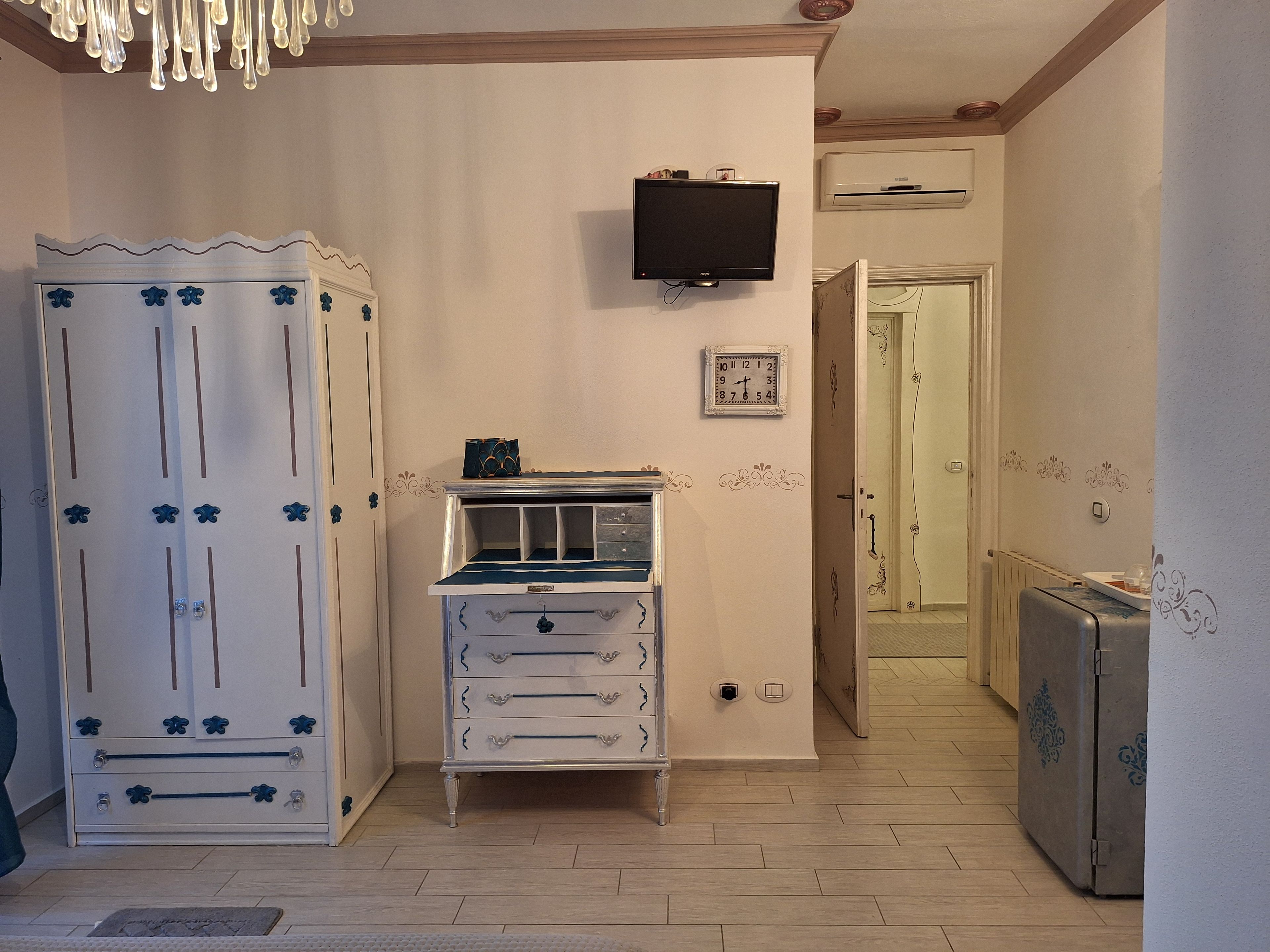 A room with a white wardrobe, a small dresser, a television mounted on the wall, and a door leading to another room.