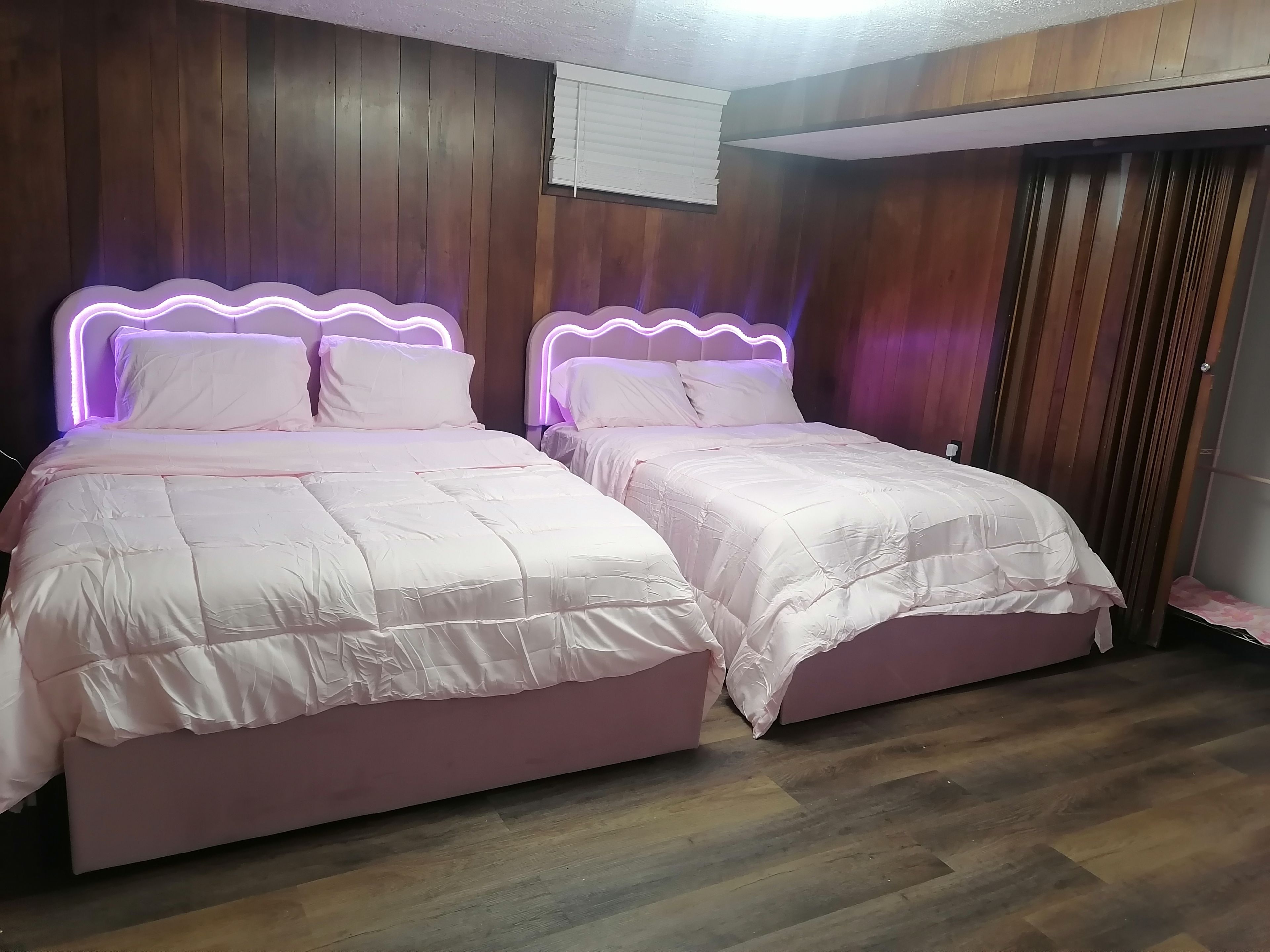 Une chambre avec deux lits, des murs en bois, un luminaire au plafond et une fenêtre avec des stores.