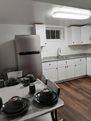 A kitchen with white cabinets, a stainless steel refrigerator, a granite countertop, and a dining table set with black dishes and teapots.