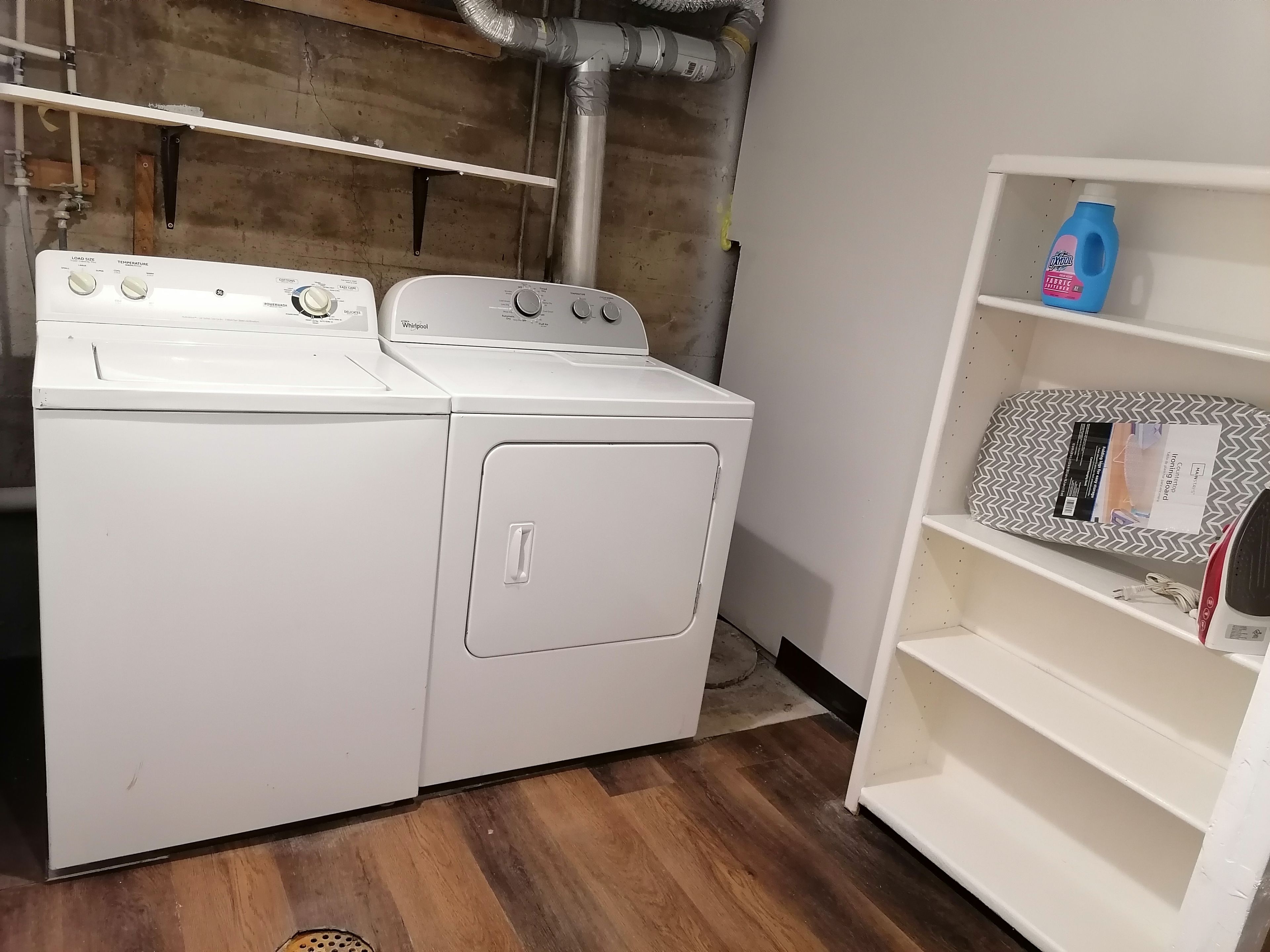 A laundry area with a stacked washer and dryer, a shelf with a detergent bottle, and a patterned box on the shelf.