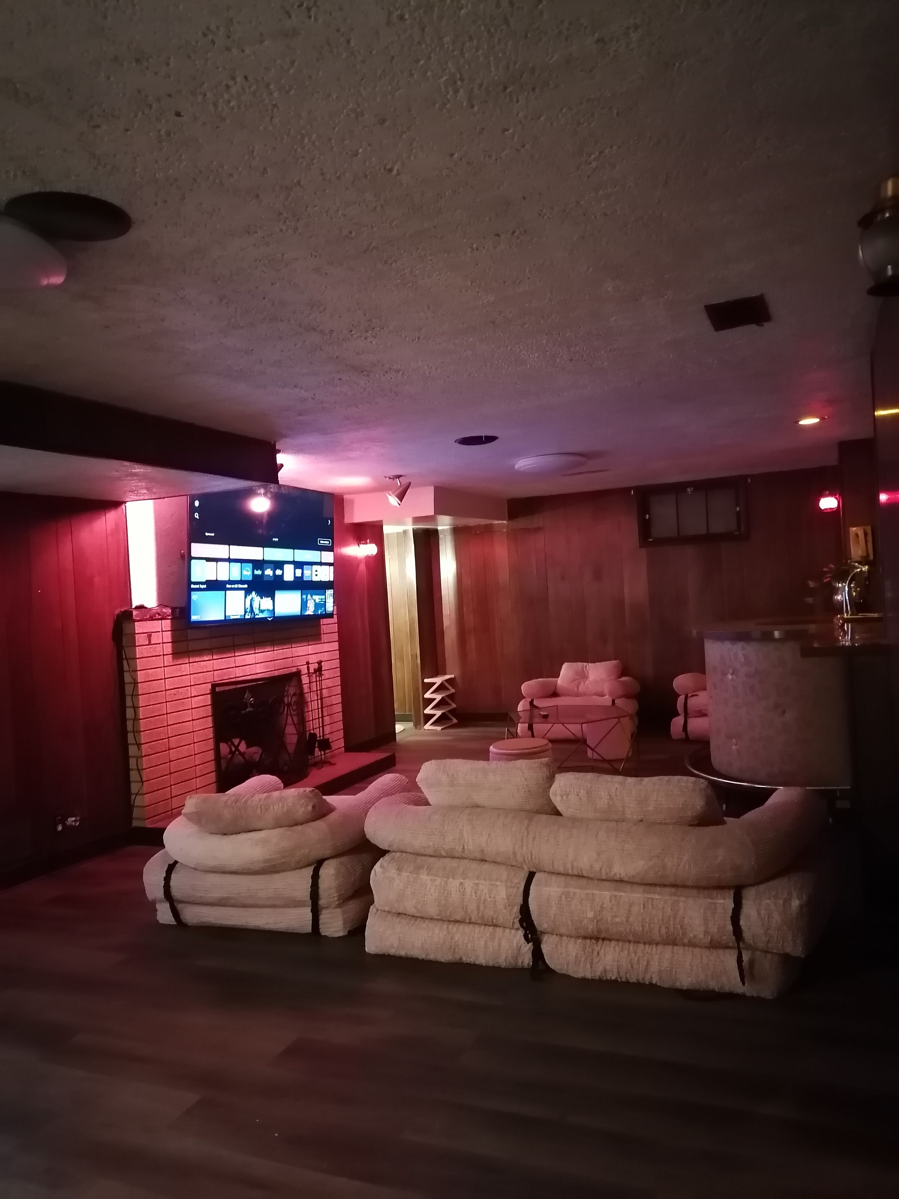 A room with a brick fireplace, a flat-screen TV mounted on the wall, and a stack of white cushions.