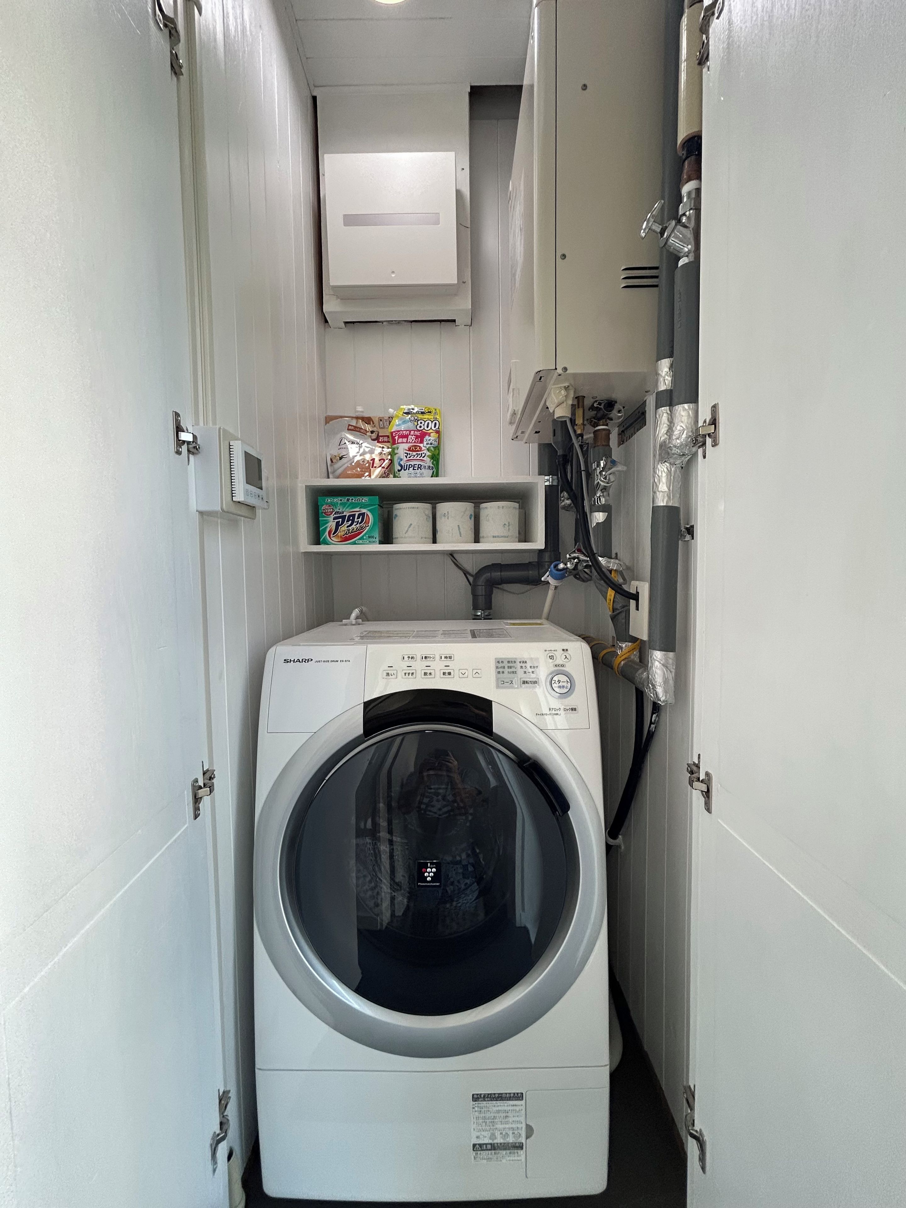 Lavanderia compacta com máquina de lavar de carregamento frontal, prateleira com produtos de limpeza e medidor de utilidades acima da máquina.