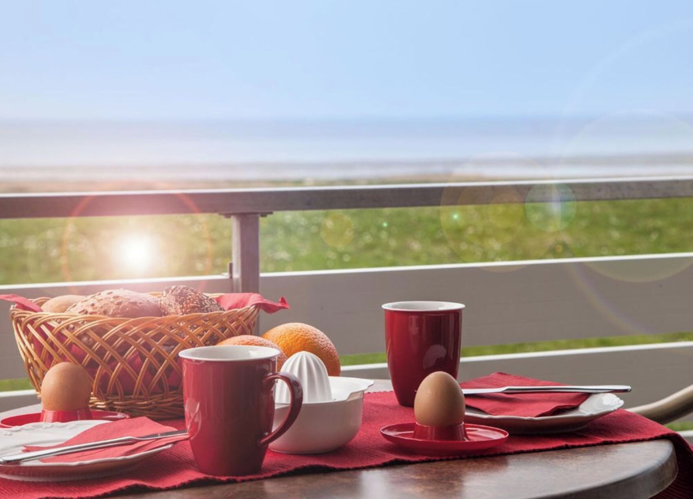 A breakfast setup with a basket of bread, a bowl, a juicer, and two mugs on a table with a red placemat, overlooking a scenic view.