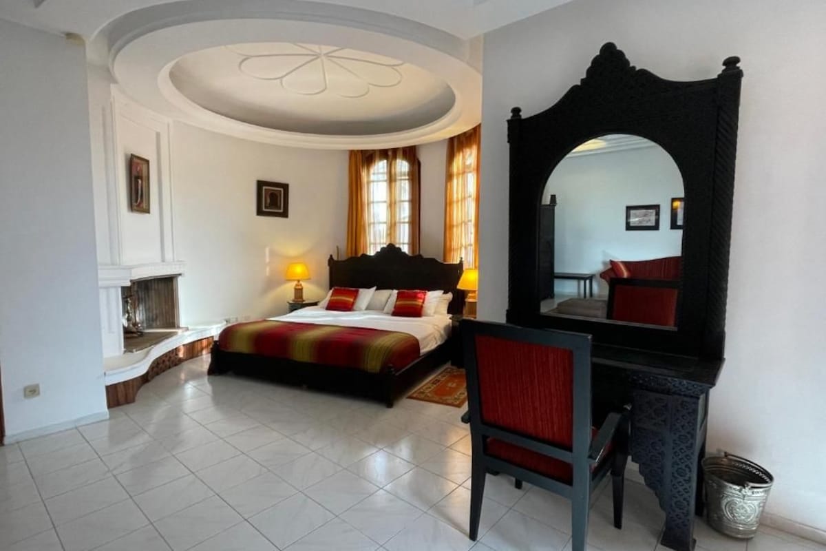 A bedroom with a bed, a desk, a chair, a fireplace, and a large mirror.