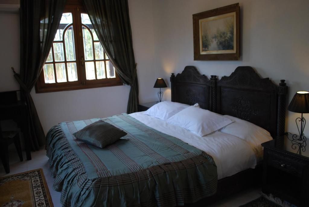 A bedroom with a bed, a nightstand, a lamp, a window with curtains, and a painting on the wall.