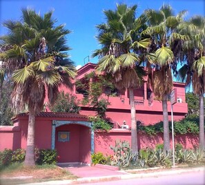 Un edificio rojo con una entrada pequeña, rodeado de palmeras y cactus.